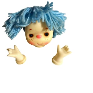 May include: A vintage doll head with blue yarn hair, rosy cheeks, freckles, and large eyes. The doll has two outstretched hands with a light skin tone. The doll's head and hands are the only visible parts.