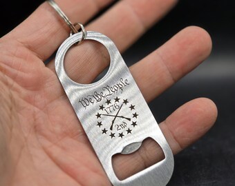 May include: A silver bottle opener with a keychain ring. The opener has an engraved design with the text "We the People 1776 2nd" and a circle of stars surrounding crossed rifles.