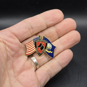 May include: A gold-colored pin with a red, white, and blue American flag on the left and a blue flag with a white star on the right. The center of the pin features a black silhouette of a dog with a bone in its mouth and the text "Military Working Dog Guardians of the Night".