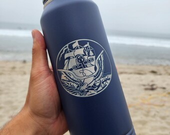 May include: A navy blue stainless steel water bottle with a silver lid and a silver base. The bottle has a white engraved design of an eagle with a ship and anchor inside a circle. The bottle is being held by a hand in front of a beach.