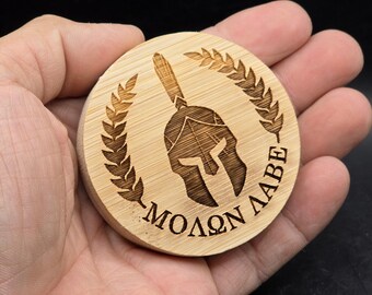 May include: A round wooden coaster with a laser-engraved Spartan helmet and laurel wreath. The text "MOLON LABE" is engraved below the helmet.