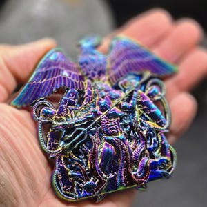 Pode incluir: Uma escultura metálica iridescente detalhada de uma águia com as asas abertas. A obra de arte apresenta detalhes intrincados e uma paleta de cores vibrantes de azuis, roxos e verdes. A escultura tem uma superfície texturizada e um design artístico único.