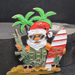 May include: A novelty Christmas ornament featuring Santa Claus in sunglasses, a military vest, and a red Santa hat, holding a rifle and a surfboard with "Merry Christmas" text. Palm trees and pineapples with sunglasses complete the tropical scene.