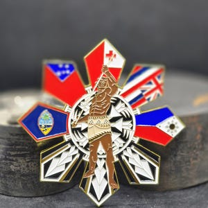 Puede incluir: Un pin detallado y multicolor que presenta una figura central sosteniendo una lanza, rodeada de banderas y formas geométricas. El pin tiene un borde dorado y un fondo oscuro. Las banderas incluyen rojo, blanco, azul y otros colores.