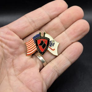 以下が含まれることがあります： 片側に赤白青のアメリカ国旗、反対側に青い十字架の白い旗が付いた金色のピン。ピンの中央には、黒い犬のシルエットが描かれた赤い盾と、「Military Working Dog, Guardians of the Night」の文字が刻まれています。