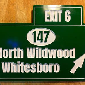 May include: A green road sign with white lettering and a white arrow. The sign reads "EXIT 6" at the top, with "147" in an oval, and "North Wildwood Whitesboro" below. The arrow points right.