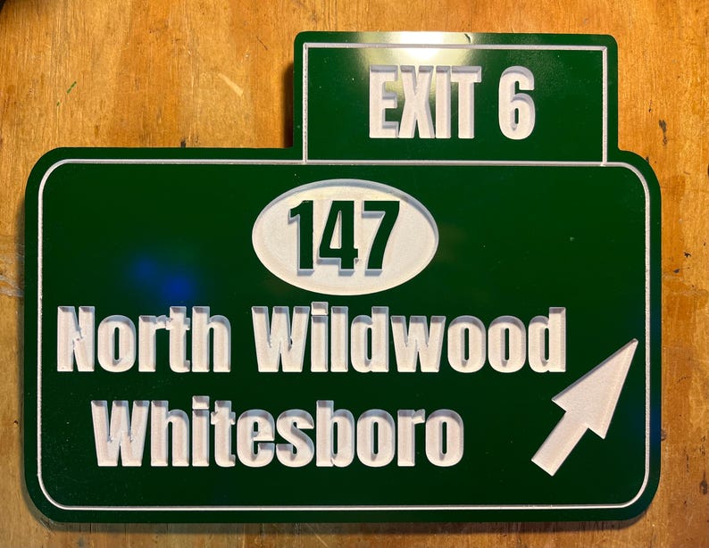 May include: A green road sign with white lettering and a white arrow. The sign reads "EXIT 6" at the top, with "147" in an oval, and "North Wildwood Whitesboro" below. The arrow points right.