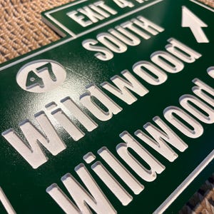 Puede incluir: Se&ntilde;al de salida de Wildwood verde y blanca. El letrero dice "EXIT 4A SOUTH Wildwood" con una flecha blanca que apunta hacia la derecha y el n&uacute;mero "47" en un c&iacute;rculo. El letrero est&aacute; hecho de un material verde oscuro con letras blancas.