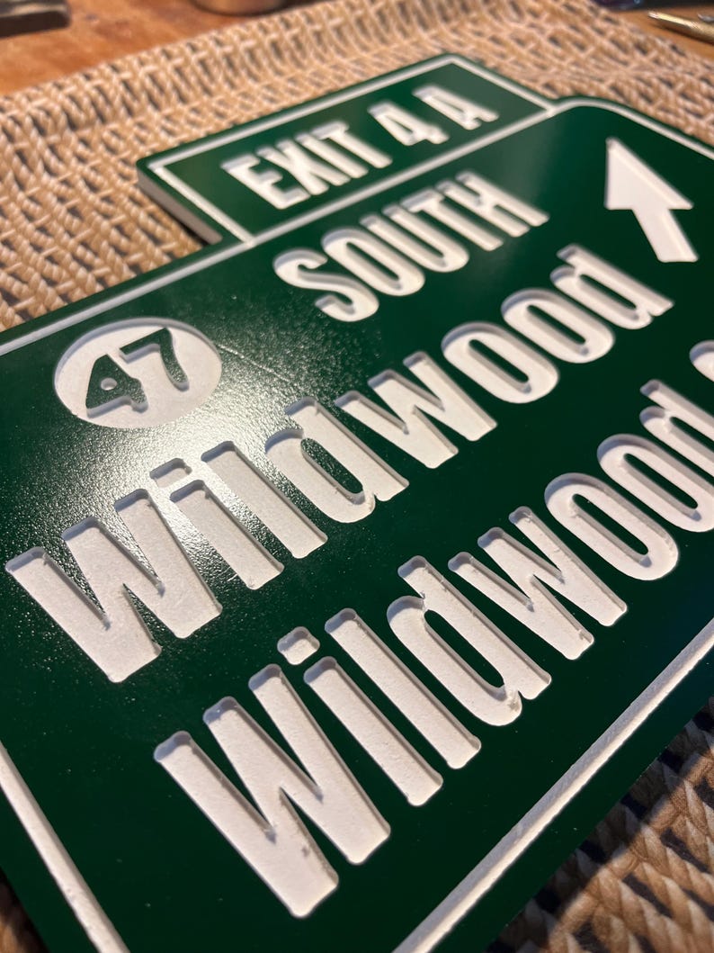 Puede incluir: Se&ntilde;al de salida de Wildwood verde y blanca. El letrero dice "EXIT 4A SOUTH Wildwood" con una flecha blanca que apunta hacia la derecha y el n&uacute;mero "47" en un c&iacute;rculo. El letrero est&aacute; hecho de un material verde oscuro con letras blancas.