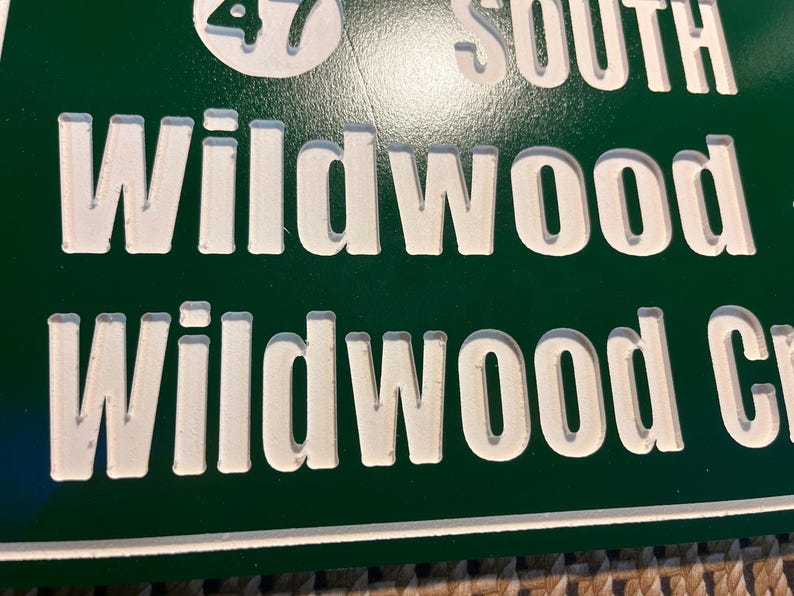 Puede incluir: Primer plano de una se&ntilde;al de tr&aacute;fico verde con letras blancas en relieve. El letrero dice "Wildwood" y "Wildwood Cr". El n&uacute;mero "47" es visible en un c&iacute;rculo blanco en la parte superior.