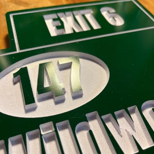 May include: Green and white sign with the words "EXIT 6" and "147" in white, and the word "WILDWOOD" in white. The sign is made of a dark green material with the text carved out.