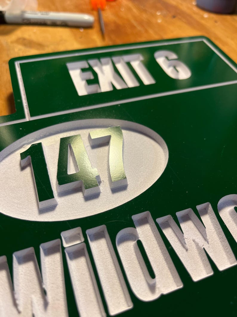 May include: Green and white sign with the words "EXIT 6" and "147" in white, and the word "WILDWOOD" in white. The sign is made of a dark green material with the text carved out.