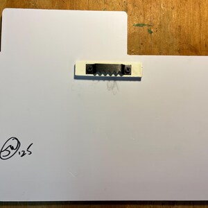 May include: A white rectangular sign with rounded corners and a small rectangular extension at the top. A metal sawtooth hanger is attached to the back. The artist's signature and the number 125 are in the lower left corner.