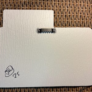 Puede incluir: Parte trasera de un letrero rectangular blanco y texturizado con una forma inusual. Un colgador de metal en forma de diente de sierra est&aacute; fijado en la parte superior. Una firma negra y el n&uacute;mero "25" est&aacute;n en la esquina inferior izquierda.