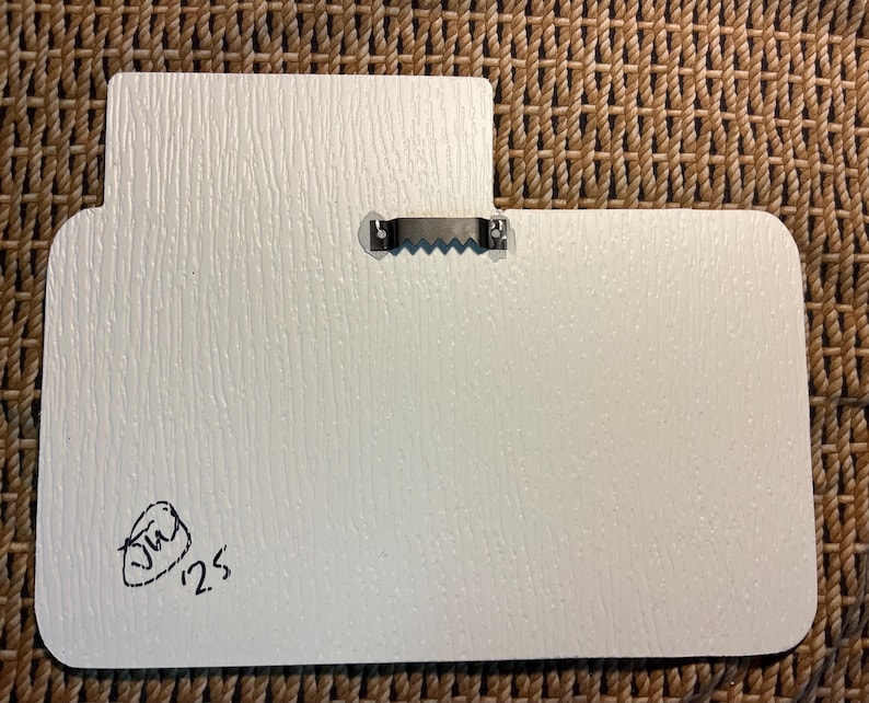 Puede incluir: Parte trasera de un letrero rectangular blanco y texturizado con una forma inusual. Un colgador de metal en forma de diente de sierra est&aacute; fijado en la parte superior. Una firma negra y el n&uacute;mero "25" est&aacute;n en la esquina inferior izquierda.