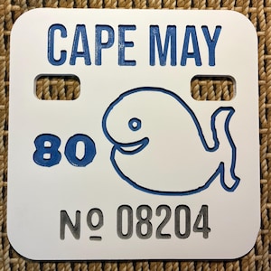 May include: White square sign with rounded corners, featuring the text "CAPE MAY" in blue, a blue whale illustration, the number "80" in blue, and the text "NO 08204" in gray.