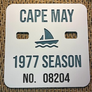 May include: White sign with rounded corners featuring the words "CAPE MAY" and "1977 SEASON" in dark blue, along with a sailboat graphic. The sign also includes the number "08204".