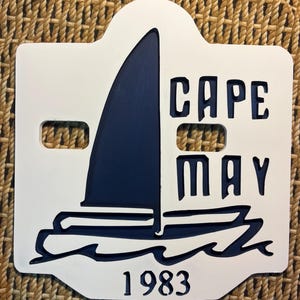 May include: A white wooden sign with a navy blue sailboat design and the words "CAPE MAY" cut out. The year "1983" is at the bottom. The sign has a decorative shape with two cut-out holes.