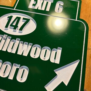May include: A green and white road sign with the text "EXIT 6" at the top. Below, the number "147" is in a white oval. The sign also includes the words "Wildwood" and "boro" with a white arrow pointing right.