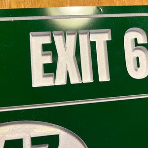 May include: Green metal sign with the white text "EXIT 6". The sign has a white border and is mounted on a wooden surface. The sign is likely used for wayfinding or directional purposes.
