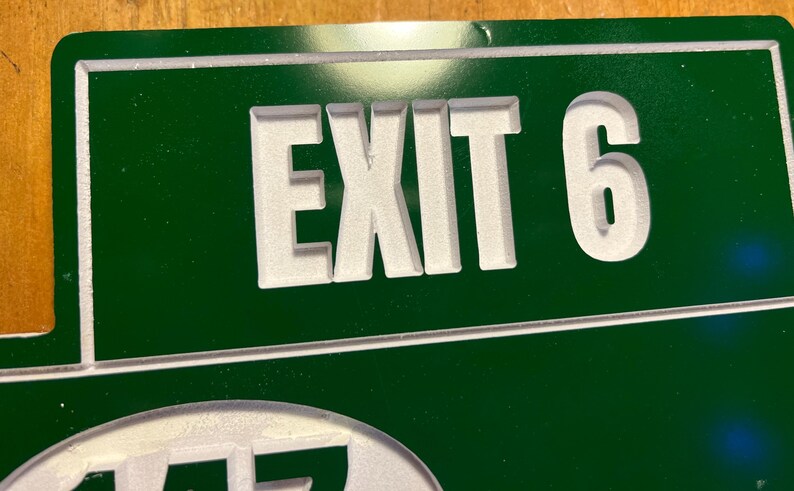 May include: Green metal sign with the white text "EXIT 6". The sign has a white border and is mounted on a wooden surface. The sign is likely used for wayfinding or directional purposes.