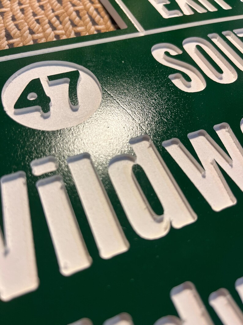 Puede incluir: Primer plano de un letrero verde con letras blancas en relieve. El letrero dice "EXIT SOUTH" y "Wildwood". Un c&iacute;rculo blanco contiene el n&uacute;mero "47". El letrero tiene una superficie texturizada.