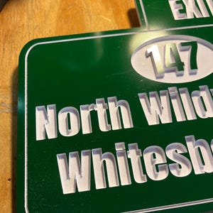 May include: A green and white road sign with the words "North Wilkesboro" and "Exit 147" in white lettering. The sign has a white border and is made of a glossy material.