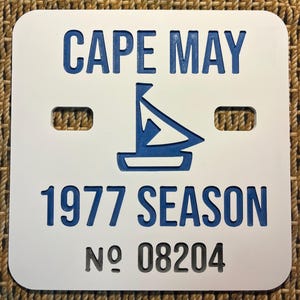 May include: White rectangular sign with rounded corners. The sign features the words "CAPE MAY" and "1977 SEASON" in blue, along with a blue sailboat graphic. The number "08204" is in black.