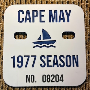 May include: White square sign with rounded corners. The sign has blue text that reads "CAPE MAY", a sailboat graphic, "1977 SEASON", and "NO. 08204". Two holes are cut out at the top.