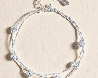 Puede incluir: Una delicada tobillera plateada con un diseño de doble cadena. La tobillera está adornada con varios abalorios esféricos calados. El cierre y una pequeña etiqueta marcada "925" son visibles. La joya se muestra sobre un fondo neutro.