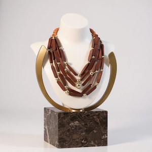 Puede incluir: Collar de varias hebras exhibido en un busto blanco. El collar presenta cuentas rectangulares de madera marrón rojizo intercaladas con pequeñas cuentas doradas. El busto está sobre una base con vetas de mármol, con un marco de metal dorado.