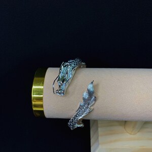Close-up of a 925 sterling silver open cuff bracelet with two intricately detailed white dragon heads and a lustrous finish. The adjustable bangle flexes for a secure fit, blending mythical elegance with bridal-ready style.