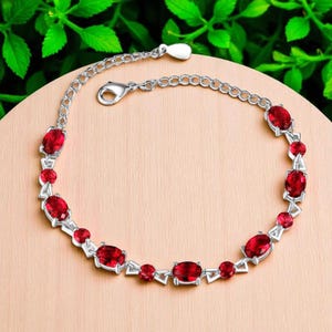 Puede incluir: Una pulsera plateada con piedras preciosas rojas ovaladas y redondas. La pulsera presenta un diseño de cadena con cierre de mosquetón. Las piedras preciosas están engastadas en un engaste de metal plateado. La pulsera se muestra sobre una superficie marrón claro.