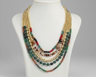 Puede incluir: Collar de múltiples hilos exhibido en un busto blanco. El collar presenta hilos de cuentas doradas, cuentas verdes, rojas, negras y marrones, y astillas de piedra natural. El collar es una pieza llamativa.