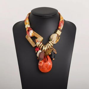 Può includere: Una collana vistosa con una catena in tessuto e dorata. La collana presenta un grande pendente arancione e un gruppo di perline colorate. La catena in tessuto ha un motivo rosso, giallo e bianco. La collana è esposta su un manichino nero.