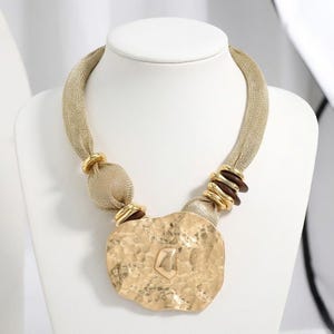 May include: A gold-toned statement necklace on a white mannequin. The necklace has a mesh chain, gold rings, and a large, textured pendant. The pendant has a hammered finish and a geometric design.