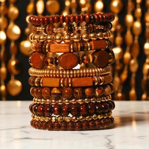 Könnte beinhalten: Ein Stapel verschiedener Armbänder in Braun- und Goldtönen. Die Armbänder bestehen aus Holzperlen, goldfarbenen Metallperlen und verschiedenen Formen. Die Armbänder sind auf einer weißen Marmoroberfläche gestapelt, mit einem verschwommenen Hintergrund aus goldfarbenen Perlen.