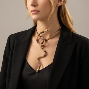 May include: A flexible snake-shaped necklace featuring a textured gold-tone mesh body with black and brown patterned segments. The serpent design wraps around the neck, with the head and tail resting vertically against the chest area.