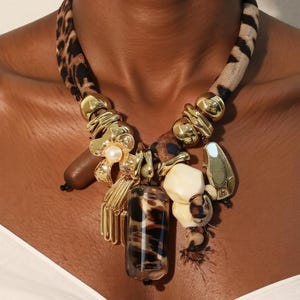 May include: A statement necklace featuring a leopard print fabric collar with gold-toned accents. The pendant includes a mix of elements: a flower with a pearl, a brown wooden piece, and a clear, cylindrical bead with a leopard print.
