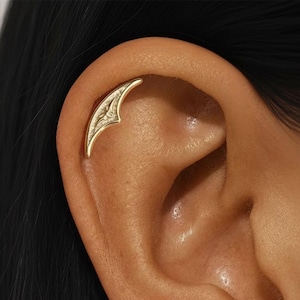 Könnte beinhalten: Ein goldfarbener Ohrschmuck mit einem blattartigen Design. Der Ohrschmuck ist gebogen, um sich der Ohrform anzupassen, und weist aufwendige Details auf. Der Schmuck wird am oberen Ohrknorpel getragen.