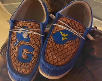 May include: Pair of blue slip-on shoes with brown leather accents. One shoe features a blue "G" and a blue silhouette. The other shoe has a blue cloud with a yellow "WV" logo. White laces and a tan sole complete the design.