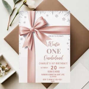 May include: A light pink invitation card with a large satin ribbon bow. The text reads "Winter ONE Onederland" and "Charlie's 1st Birthday!" with the date and location. The card is decorated with snowflakes.