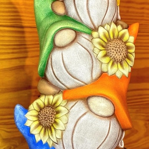 May include: A whimsical stack of three garden gnomes, each with a different colored hat: yellow, green, orange, and blue. Each gnome has a sunflower attached to its hat. The gnomes are painted in neutral tones with detailed features.