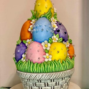 May include: A decorative Easter basket filled with brightly coloured eggs. The eggs are painted in yellow, blue, orange, pink, and purple, decorated with clear crystals and surrounded by white flowers and green grass. The basket is grey and has a woven design.