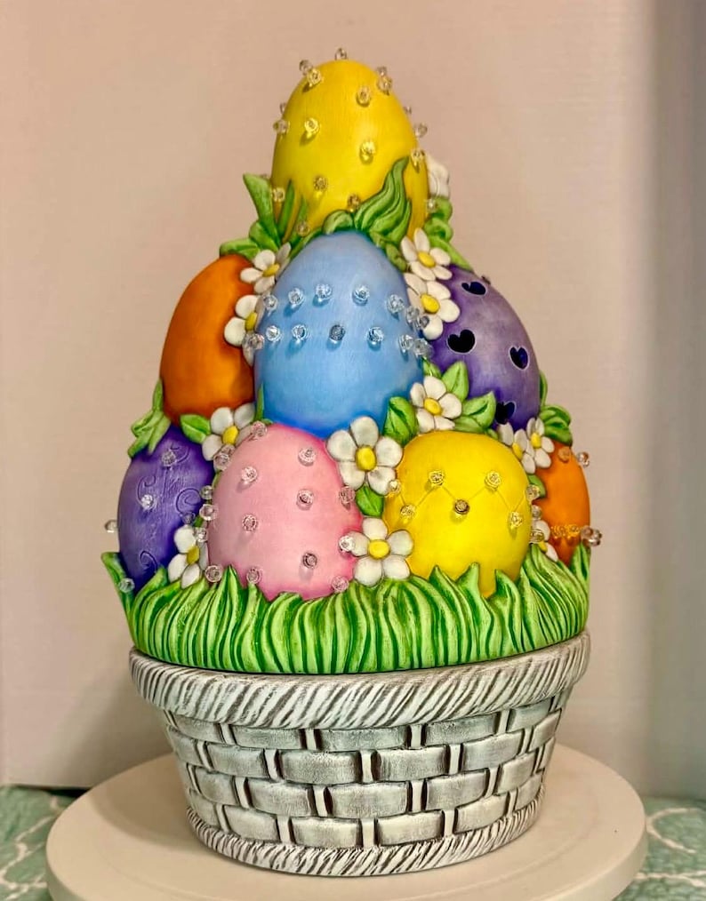 May include: A decorative Easter basket filled with brightly coloured eggs. The eggs are painted in yellow, blue, orange, pink, and purple, decorated with clear crystals and surrounded by white flowers and green grass. The basket is grey and has a woven design.