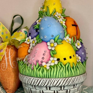 May include: A ceramic Easter basket filled with brightly coloured eggs. The eggs are decorated with white flowers and sparkling embellishments. The basket is grey with a woven pattern, and the base is surrounded by green grass.