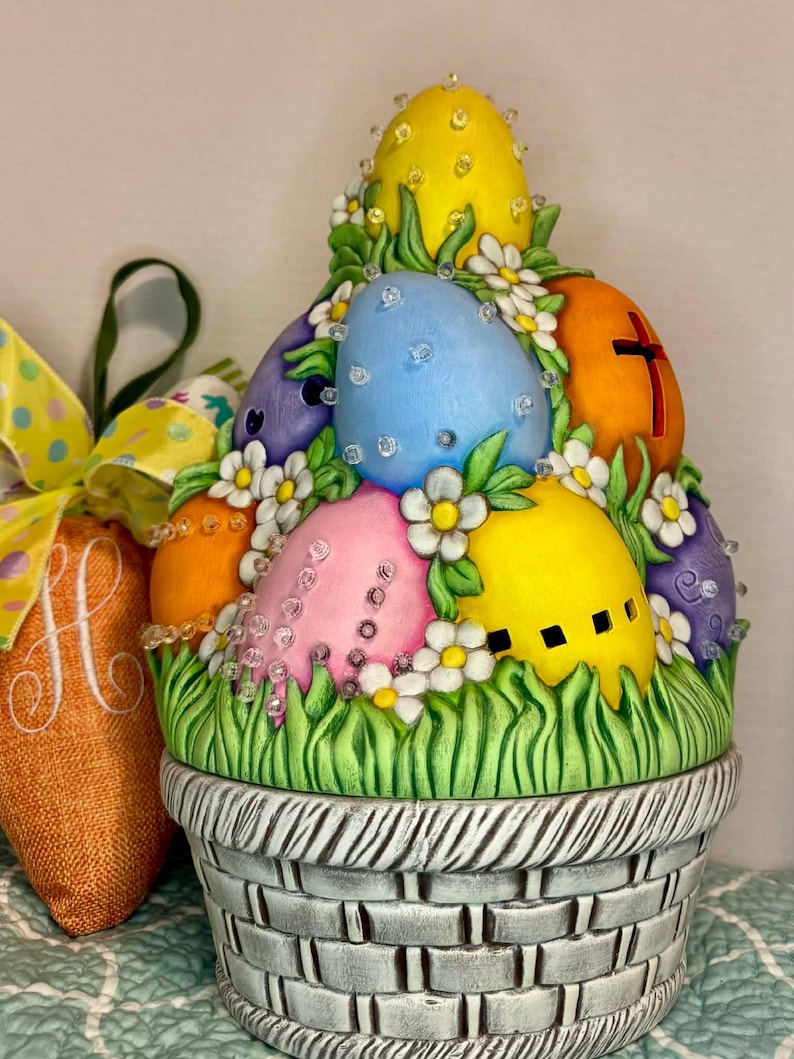 May include: A ceramic Easter basket filled with brightly coloured eggs. The eggs are decorated with white flowers and sparkling embellishments. The basket is grey with a woven pattern, and the base is surrounded by green grass.