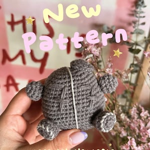 Puede incluir: Un pequeño juguete de peluche tejido a ganchillo en color gris oscuro, de forma redonda y superficie texturizada, con detalles de cruces cosidas en el frente, una línea vertical de hilo blanco y pequeñas protuberancias. El texto New Pattern +appreciationtester es visible.