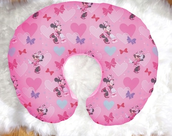 Op de afbeelding: Een roze voedingskussen met een herhalend patroon van Minnie Mouse, harten en strikken. Het kussen is ontworpen met een gebogen vorm voor comfortabele ondersteuning. Het patroon bevat verschillende tinten roze, rood en paars.