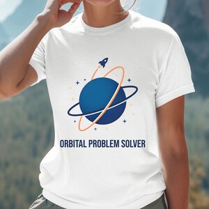 Peut inclure: T-shirt blanc avec un motif de planète bleue avec des anneaux et une fusée, et le texte "ORBITAL PROBLEM SOLVER". Le t-shirt est de style col rond.